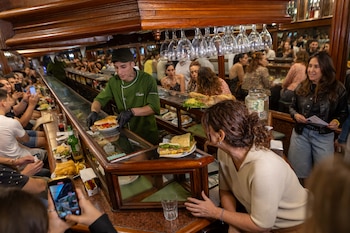 Un camarero con uniforme verde y guantes negros sirve sándwiches en una barra de madera con exhibiciones de comida y copas colgadas. Hay clientes y teléfonos móviles
