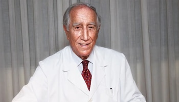 Retrato de primer plano de Enrique Moreno González, un hombre mayor con pelo gris, bata blanca, camisa de rayas y corbata roja, sonriendo