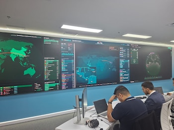 Dos hombres sentados frente a computadoras portátiles ante una gran pantalla mural que proyecta mapas mundiales y gráficos con datos