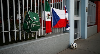 Mochila escolar verde, bufanda con colores de México, bandera de Chequia, reja metálica, muro de escuela azul y rojo, balón de fútbol en acera.