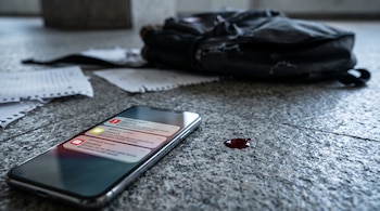 Teléfono sobre un piso de granito con notificaciones en pantalla, una gota roja al lado y una mochila con hojas de papel al fondo.