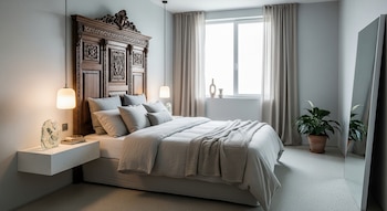 Dormitorio luminoso con paredes grises, cama con cabecero de madera tallada, ropa de cama gris, dos lámparas colgantes, mesita flotante y ventana.