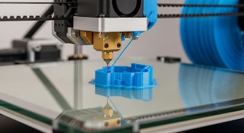 Primer plano de una impresora 3D. La boquilla extrude filamento azul para construir un objeto en una base reflectante. Carrete azul al fondo.