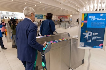 Hombre de traje azul usando un teléfono móvil en un torniquete. Un cartel con texto "tapp PILOT Stations" y un código QR. Varias personas