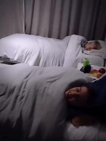 Imagen de dos niños en camas individuales con ropa de cama blanca. Un niño rubio duerme tranquilamente, mientras otro niño de cabello oscuro sonríe a la cámara