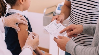 Primer plano de una niña recibiendo una inyección en el brazo por una mano enguantada. Manos de adultos sostienen un carné de vacunación.
