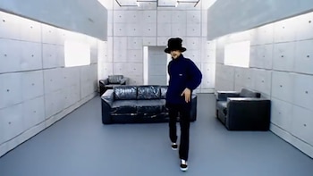 Jamiroquai, con sombrero oscuro y ropa azul y negra, caminando en una habitación gris con paredes de concreto y sofás negros