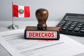 Sello de madera con la palabra DERECHOS en rojo, un documento, una bandera de Perú y una calculadora sobre una superficie gris clara.