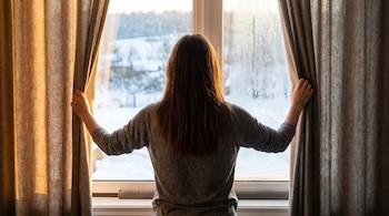 Una persona de espaldas, con cabello castaño, abre unas cortinas de color beige-marrón para ver a través de una ventana con condensación y un paisaje nevado al exterior.
