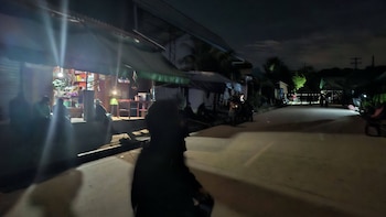 Vista nocturna de una calle oscura en una comunidad, con personas sentadas y de pie, y una tienda con luz brillante al fondo. El cielo está nublado
