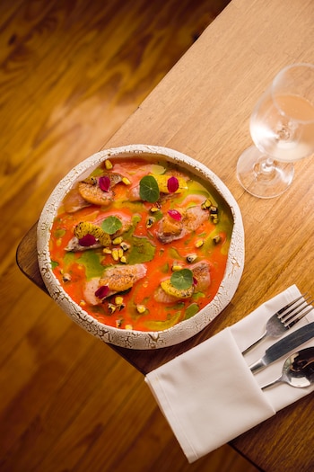 Un plato gourmet en un cuenco texturizado, con salsa roja y verde, pescado crudo, rodajas de naranja quemada, maíz y hierbas, junto a una copa de vino y cubiertos en mesa de madera clara