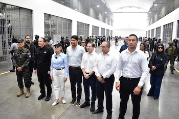 Grupo de personas con ropa formal y uniformes militares en el pasillo de una prisión flanqueado por celdas con rejas y personas en el fondo