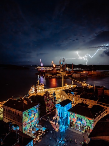 Vista aérea nocturna de edificios con proyecciones de colores, un puerto con barcos y un rayo en el cielo
