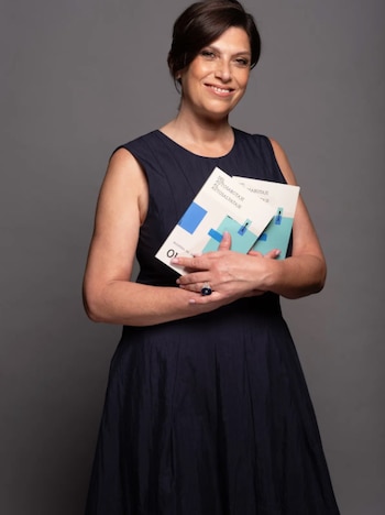 Mujer con cabello oscuro y sonrisa posa frente a un fondo gris, sosteniendo dos libros con las portadas visibles, que muestran el título 'DEL AUTOSABOTAJE AL AUTOSALVATAJE'