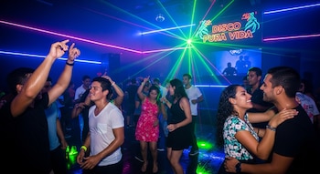 Grupo de personas bailando en una discoteca con luces de neón azules, rosadas y láseres verdes. Se ve un letrero luminoso con el texto 'DISCO PURA VIDA'.
