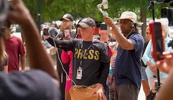 Un grupo de periodistas, algunos con camisetas que dicen 'PRESS', graba un evento al aire libre utilizando teléfonos inteligentes con micrófonos y cámaras profesionales
