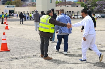Hombre con chaleco de Migración saluda a otro. Otros caminan hacia el Centro de Recepción de Retornados, con conos naranjas en un área pavimentada
