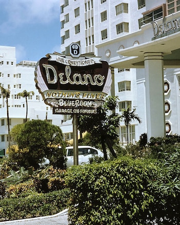 Letrero grande y retro del Delano Miami Beach Hotel con letras blancas, rodeado de vegetación tropical; la fachada blanca del hotel y un coche se ven al fondo