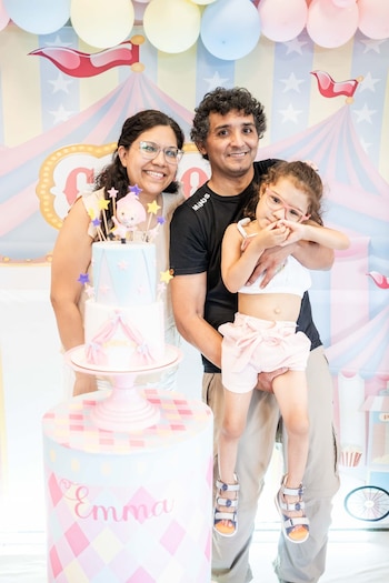 Una niña con lentes es sostenida por su padre mientras sonríe junto a su madre, con un pastel de dos pisos y un fondo de circo en su cumpleaños