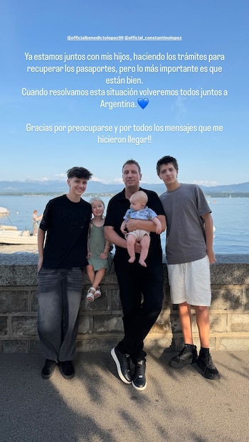 Um homem segurando um bebê, com dois meninos e uma menina, sorrindo e olhando para frente, com um lago e montanhas ao fundo sob um céu azul