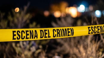 Cinta amarilla con la frase "ESCENA DEL CRIMEN" en letras negras, sobre un fondo nocturno desenfocado con vegetación y luces tenues.