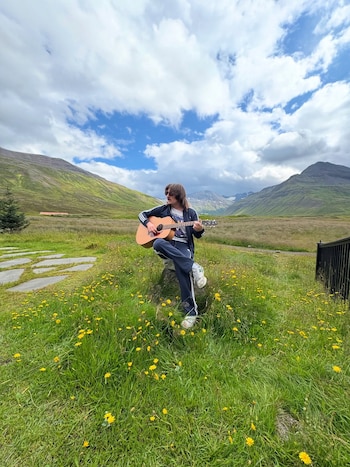 Joven con gafas y ropa deportiva toca guitarra acústica sentado en una roca. Hay un campo de hierba con flores amarillas, montañas y cielo nublado