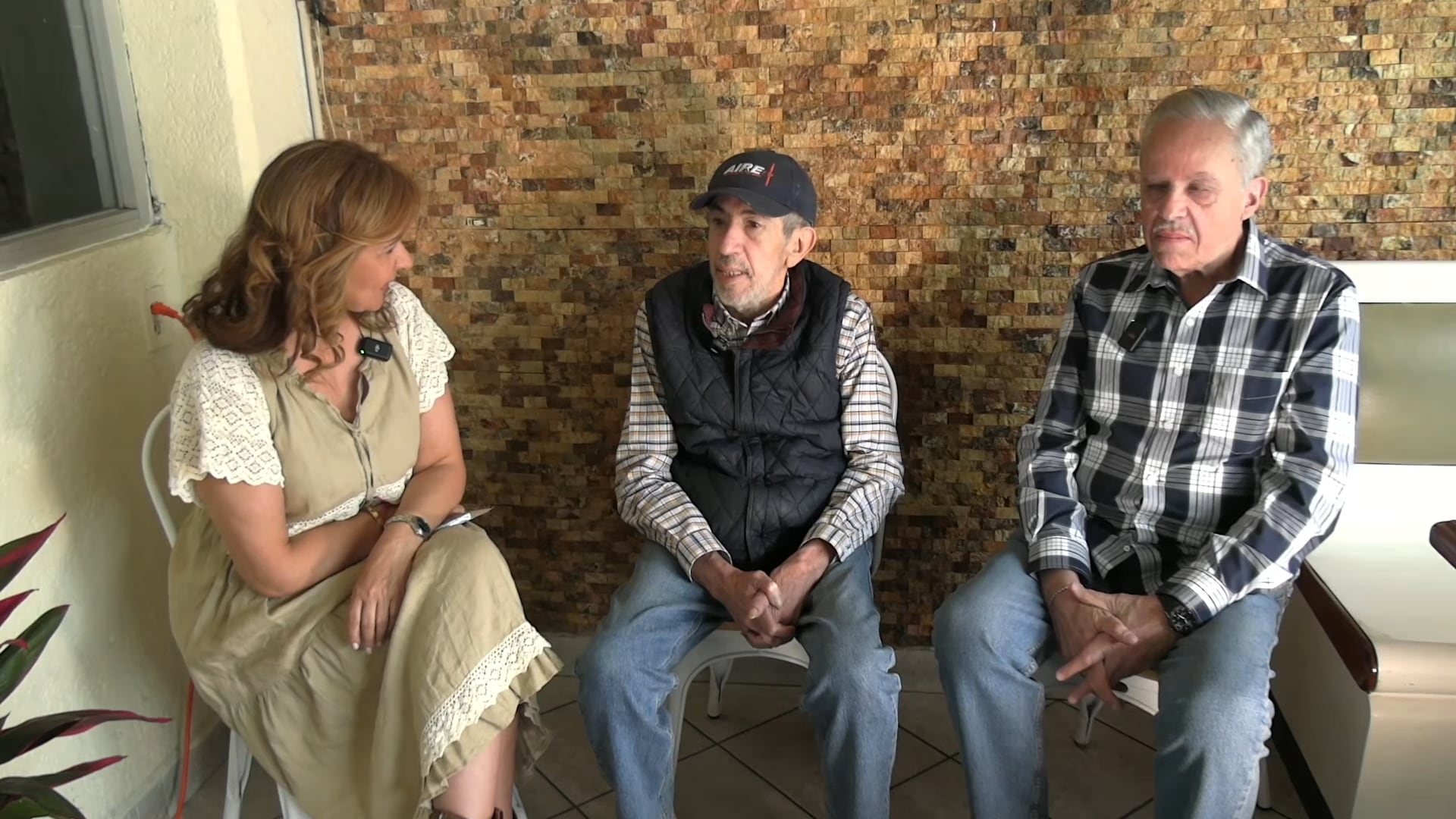La periodista Matilde Obregón entrevista al actor Ricardo Hill y a su hermano, quienes comparten una conversación sentados frente a una pared de piedra. (Captura de pantalla: YouTube)