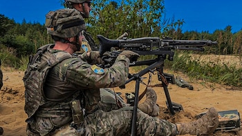 Dos soldados uniformados en camuflaje. Un soldado opera una ametralladora montada sobre trípode. Terreno arenoso con vegetación y cielo azul. Caja de municiones