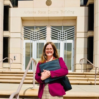 Una mujer con gafas y chaqueta magenta sostiene una carpeta negra frente a las puertas del Edificio de los Tribunales Estatales de Arizona