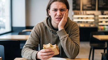 Hombre joven con sudadera caqui se toca la mandíbula con la mano, tiene una expresión de dolor y sostiene un sándwich mordido. Está sentado en un restaurante.