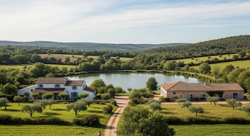 Dos casas con techos de teja, un lago, árboles, campos verdes, un camino de tierra y colinas bajo un cielo azul claro con nubes.