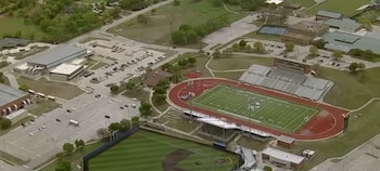 Fotografía aérea de un complejo escolar con un campo de fútbol americano, pista de atletismo, campo de béisbol, varios edificios y estacionamientos llenos de vehículos