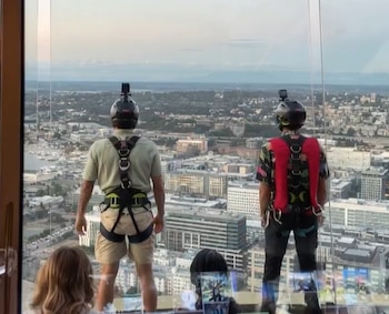 Vista trasera de dos hombres con cascos equipados con cámara y arneses, de pie frente a un ventanal con vista a la ciudad de Seattle y un puerto