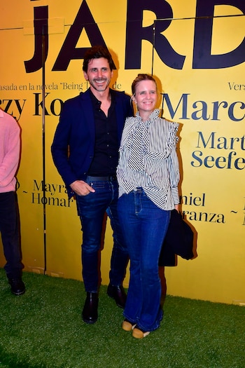 Un hombre y una mujer sonríen frente a un fondo amarillo con letras grandes. Él lleva chaqueta azul, camisa negra y vaqueros; ella, blusa a rayas y vaqueros. Ambos están de pie sobre césped artificial