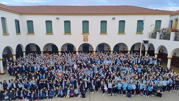 Cientos de niños con uniforme azul y celeste levantan las manos en el patio de una escuela frente a una fachada blanca con arcos y techo de tejas