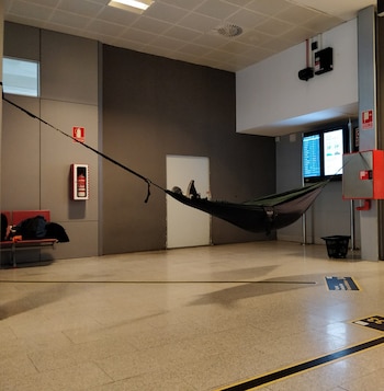 Una hamaca verde con pies de una persona suspendida entre dos columnas en un aeropuerto, con un extintor, bancos, una mochila y pantallas de información