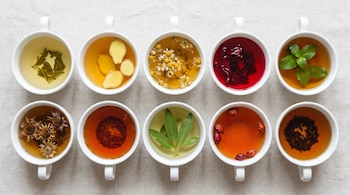 Diez tazas blancas con diferentes infusiones y tés de variados colores y texturas de ingredientes, dispuestas en dos filas sobre una superficie de tela clara, vista cenital.