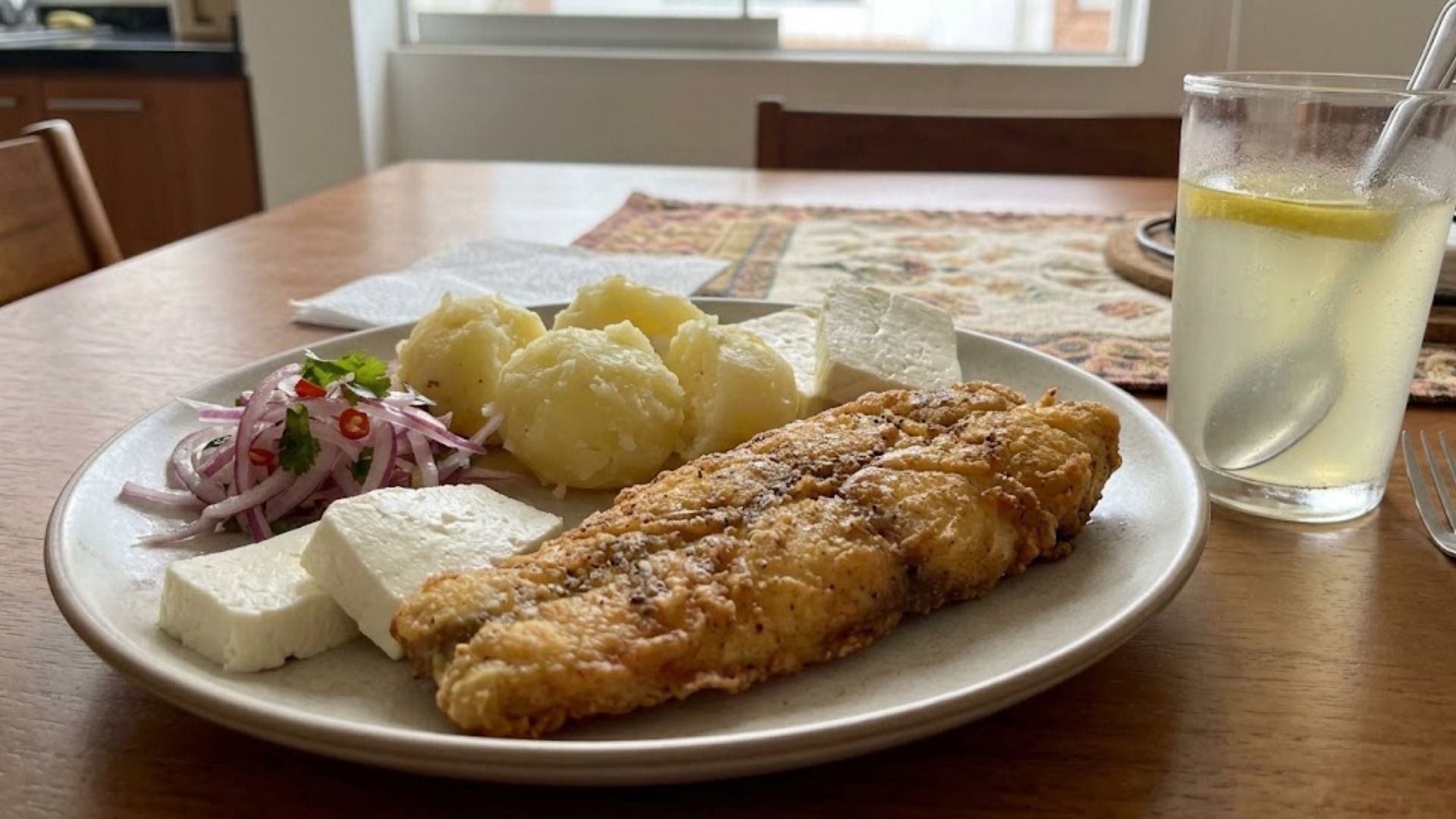 Un plato servido con milanesa frita, papas cocidas, queso fresco y ensalada de cebolla se acompaña de un vaso de limonada sobre una mesa de madera. (Imagen Ilustrativa Infobae)