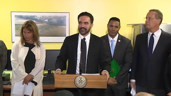 Zohran Mamdani habla en un podio de madera. A su izquierda está Rebecca Slaughter, y a su derecha Alvaro Bedoya, con otros de pie en una sala amarilla