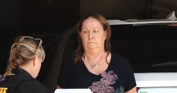 Una mujer de mediana edad con cabello castaño y blusa oscura es asistida por una agente de uniforme del sheriff con trenza rubia, cerca de un vehículo
