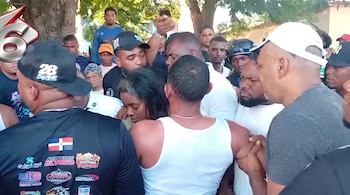 Varias personas se agrupan en una calle alrededor de una mujer y un hombre en camiseta blanca en un entorno con árboles.