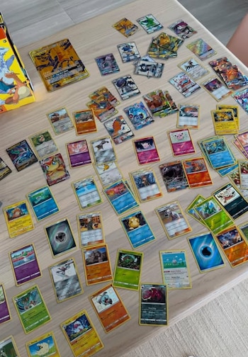 Vista aérea de decenas de cartas coleccionables Pokémon coloridas y brillantes esparcidas sobre una mesa de madera clara, junto a una caja de refuerzo