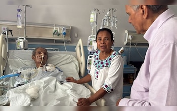 Un hombre y una mujer al lado de un paciente en una cama de hospital con tubos médicos. El paciente es un hombre de edad avanzada con un tubo nasal