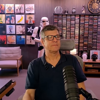 Un hombre con gafas y camiseta tipo polo se encuentra frente a un micrófono en una sala con carátulas de vinilos y estanterías de discos.