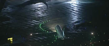 Vista aérea nocturna de un avión sobre el pavimento oscuro de una pista con hileras de luces verdes y amarillas