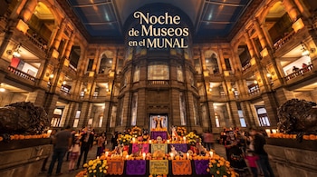 Patio interior con arcos de cantera, un altar con flores de cempasúchil, velas, personas y el texto Noche de Museos en el MUNAL.
