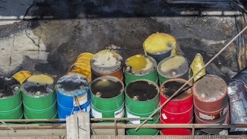 Barriles de colores verde, azul, rojo, amarillo y óxido, algunos volcados o con tapas oscuras, sobre una superficie gris y negra manchada