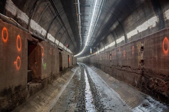 Vista interior de un túnel con paredes y techo de hormigón. Presenta marcas circulares anaranjadas, tuberías, luces, un lecho de grava y agua