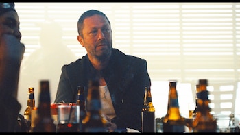 Hombre de pelo oscuro y barba corta, vestido con chaqueta oscura, sentado en una mesa. Botellas de cerveza en primer plano. Fondo claro con líneas horizontales