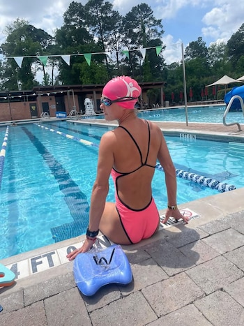 Una mujer con gorro de natación rosa y traje de baño rosa y negro sentada al borde de una piscina, con una tabla de natación azul a su lado